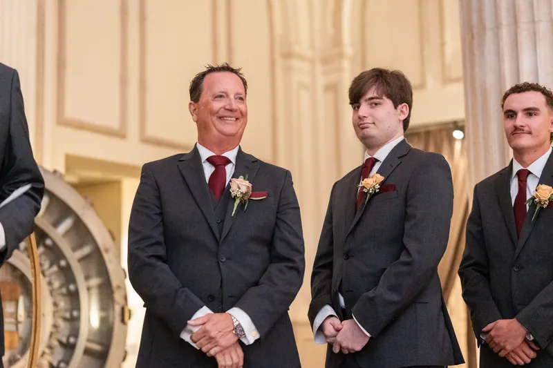 Four groomsmen in matching black suits with floral pins smile while standing near a clock fixture.