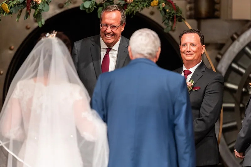 Stacy in a white lace gown and veil stands with Dean and two other men in suits beneath an arched doorway at The Treasury