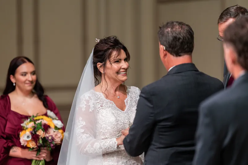 Stacy beams while speaking with Dean, wearing her lace wedding dress and veil as a guest in burgundy watches nearby.