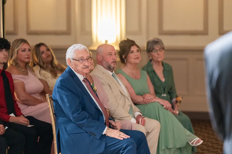 Wedding guests sit in wooden chairs facing the ceremony, including an older man in a blue jacket and woman in green.