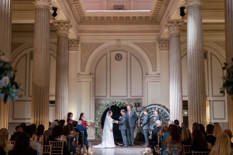The Treasury on the Plaza's neoclassical ceremony hall fills with seated guests as the couple stands before a decorated arch.