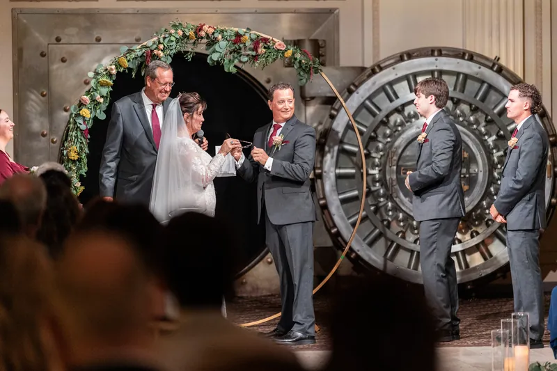 Dean stands at the floral arch laughing as Stacy approaches, surrounded by attendants in gray suits in the elegant ballroom.