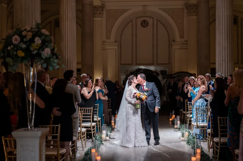 The newlyweds process through a candlelit aisle lined with guests in the venue's formal reception space.