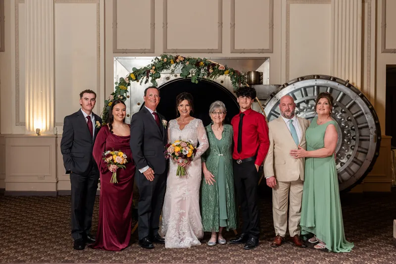 Stacy stands with five family members in formal attire, holding her bouquet in front of the large clock.