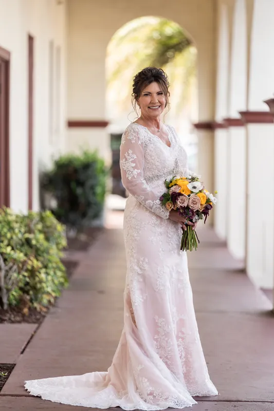 Stacy stands alone in a lace gown holding a yellow and coral bouquet beneath The Treasury on the Plaza's arched loggia.