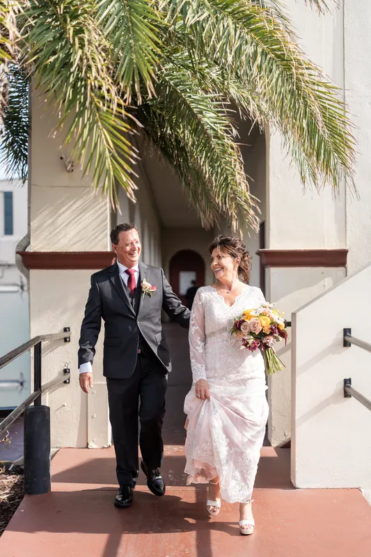 Dean and Stacy exit through palm fronds onto the terracotta portico, walking toward the entrance.