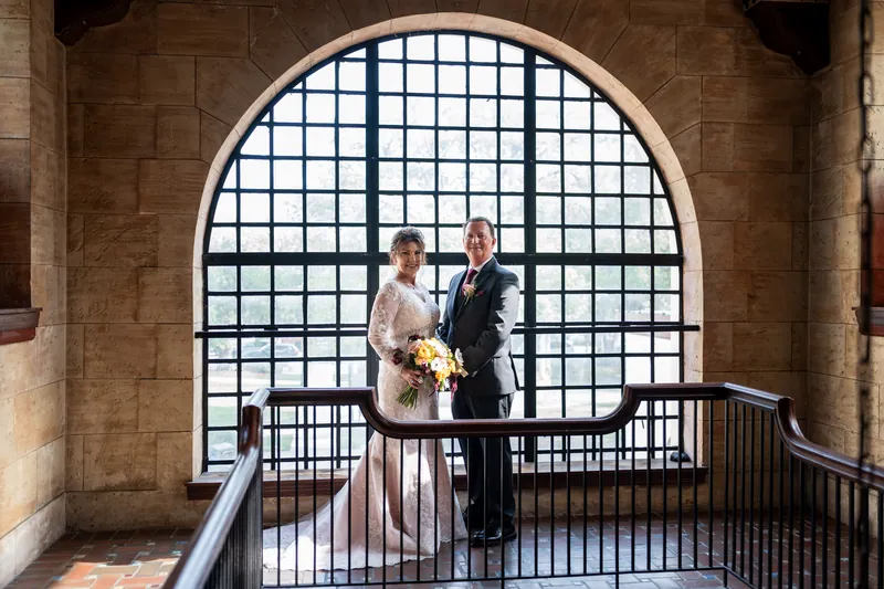 The couple stands beneath a massive arched window with black gridwork, light streaming through behind them.