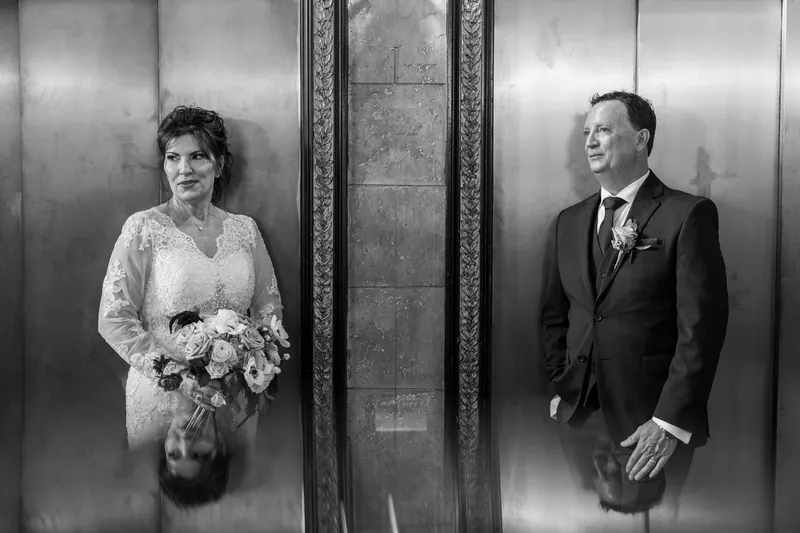 Stacy and Dean stand in an ornate elevator, Stacy holding a white bouquet in a lace gown and Dean in a dark suit