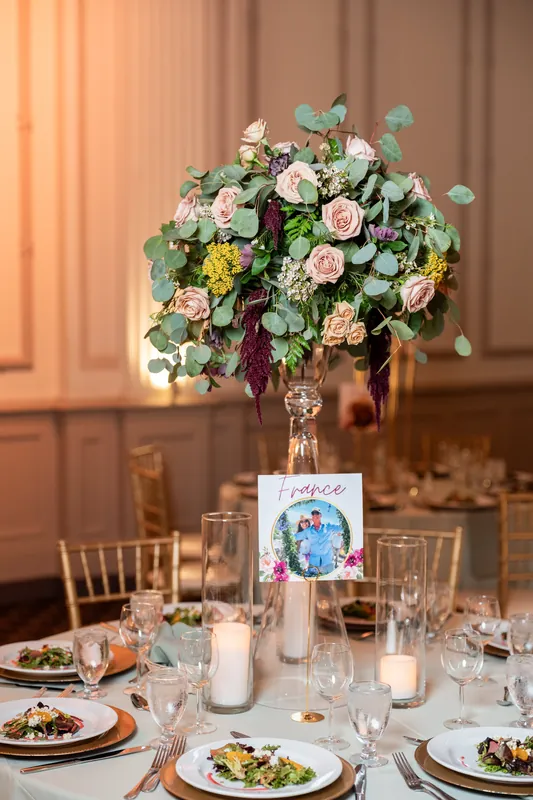 A tall arrangement of pale roses and greenery anchors a banquet table set with place settings and candles.