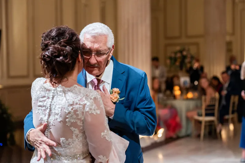 Stacy dances with an older gentleman in a blue blazer, her lace gown catching the warm architectural light of the historic