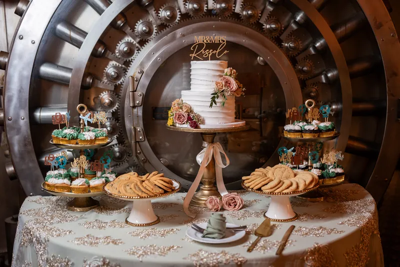 A tiered white wedding cake with floral accents sits on a pedestal inside The Treasury's ornate arched vault, flanked