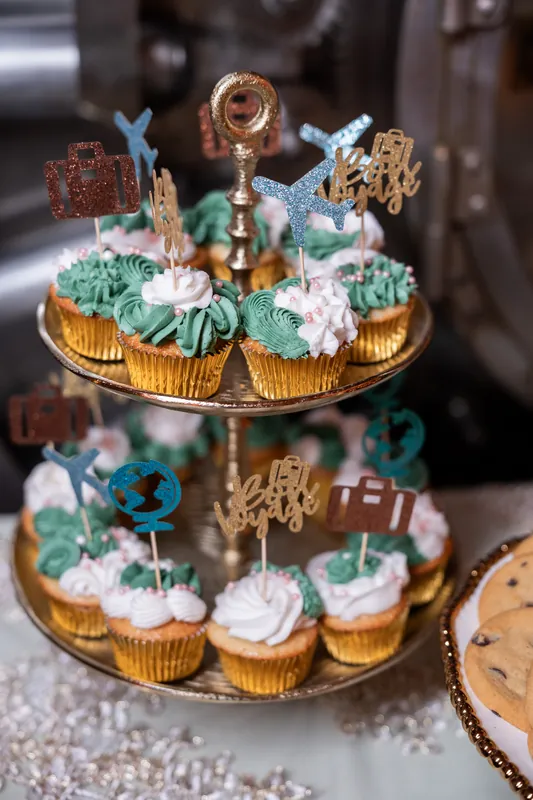 A two-tiered cupcake tower features pistachio-frosted cupcakes with gold leaf details and decorative toppers on a stone
