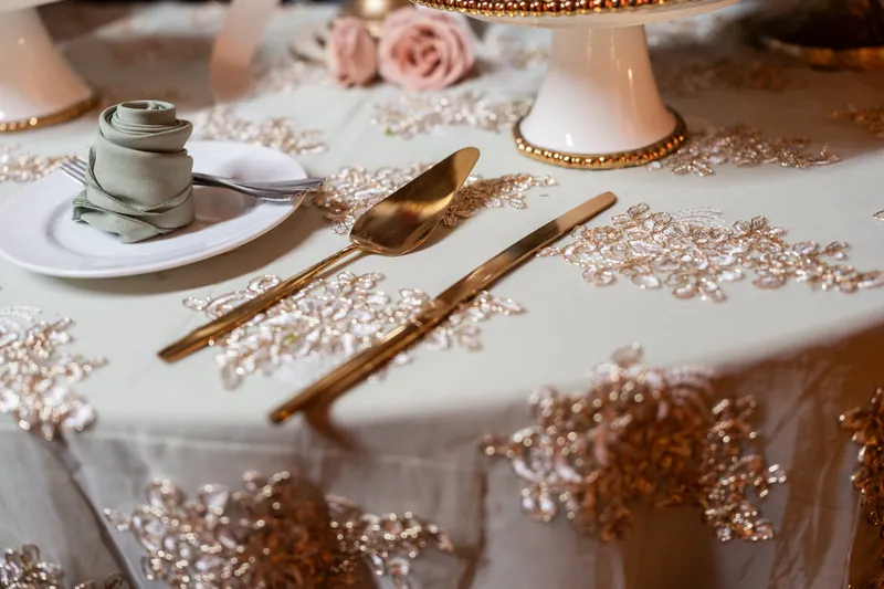 Gold flatware and a white napkin with rings rest on a cream tablecloth scattered with rose-gold beads and blush florals.