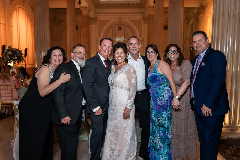 Stacy in a white lace gown stands with eight family members in formal attire inside The Treasury's classical arched hall.