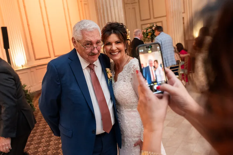 Stacy poses beside an older man in glasses and a navy suit while a guest photographs them at The Treasury on the Plaza.