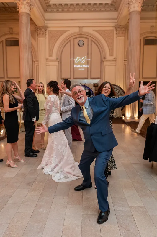 Dean celebrates with an exuberant gesture in a navy suit as Stacy stands nearby in her lace gown among reception guests.