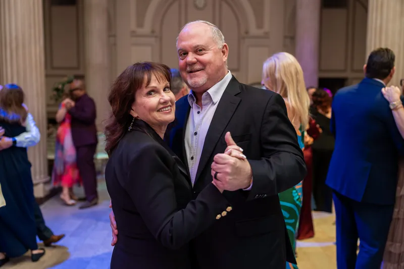 A guest in dark clothing gives a thumbs-up while dancing with a man in a blazer during the celebration.