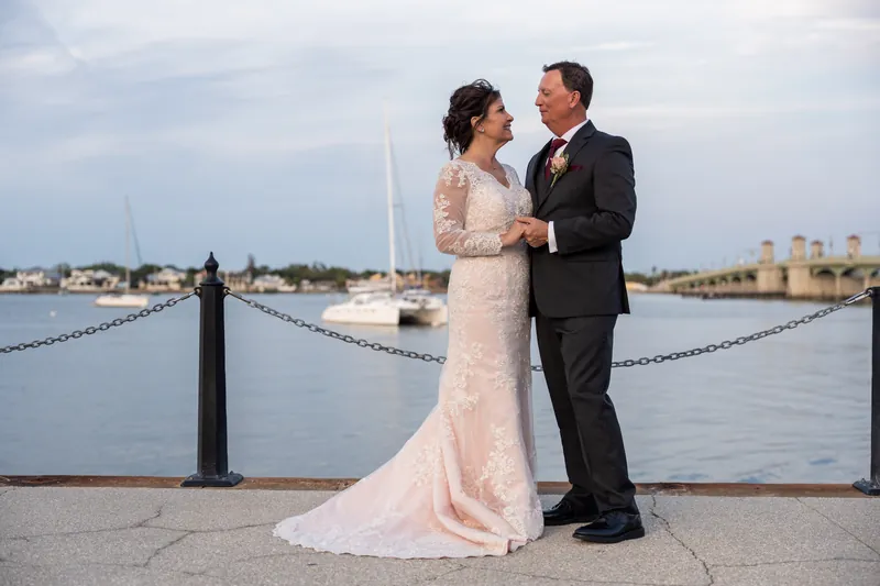 Stacy and Dean stand arm-in-arm overlooking the waterfront at St. Augustine Bayfront, sailboats visible across the water.