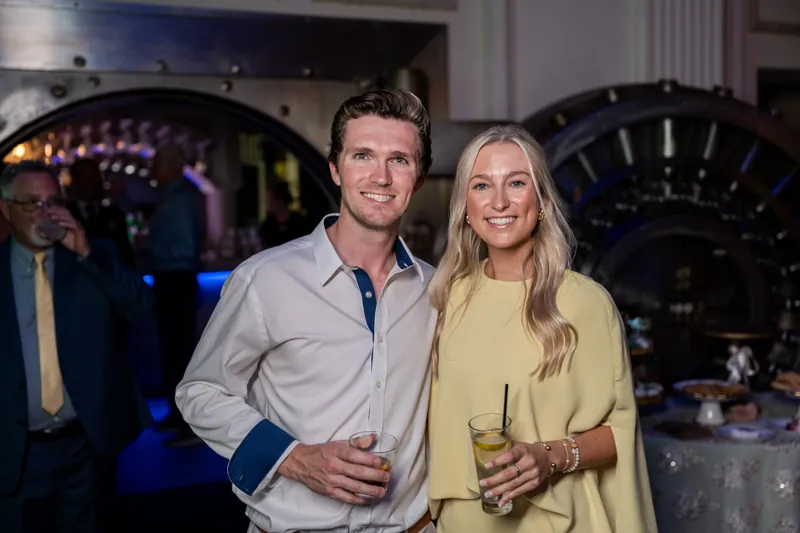 Dean and Stacy hold drinks while posing together at an indoor reception with purple architectural details behind them.