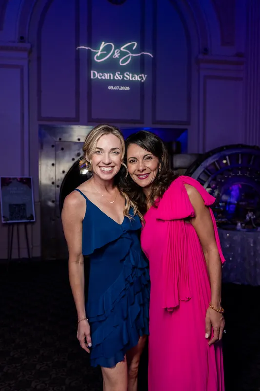 Stacy in a navy dress and a guest in hot pink stand together under the couple's monogrammed projection.