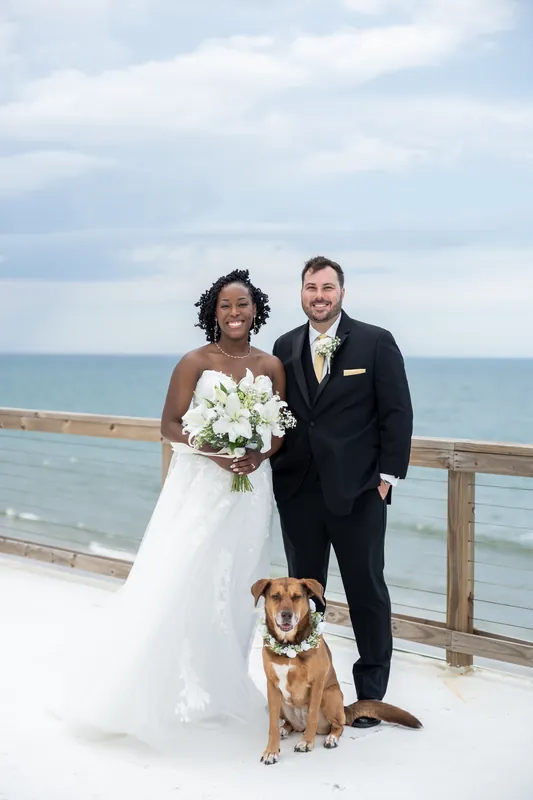 Malika in a white gown and Stephen in a dark suit stand with a brown dog on a seaside deck overlooking turquoise water.