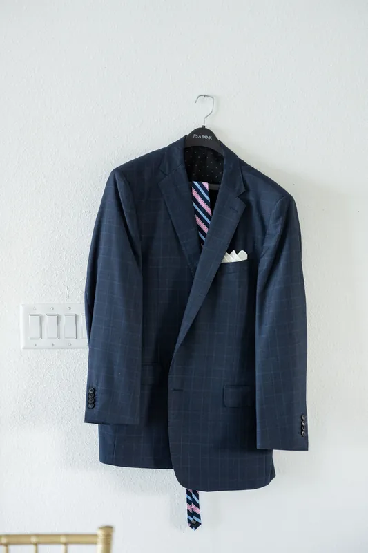 Navy blazer with striped tie draped over hanger on white wall, pocket square visible.