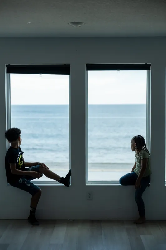 Two people sit facing each other on opposite windowsills, ocean and beach visible through large windows behind them.