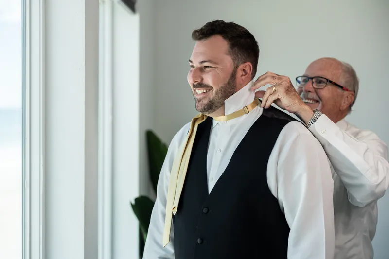 Person in white shirt and black vest smiles while another adjusts their gold tie in a bright room.