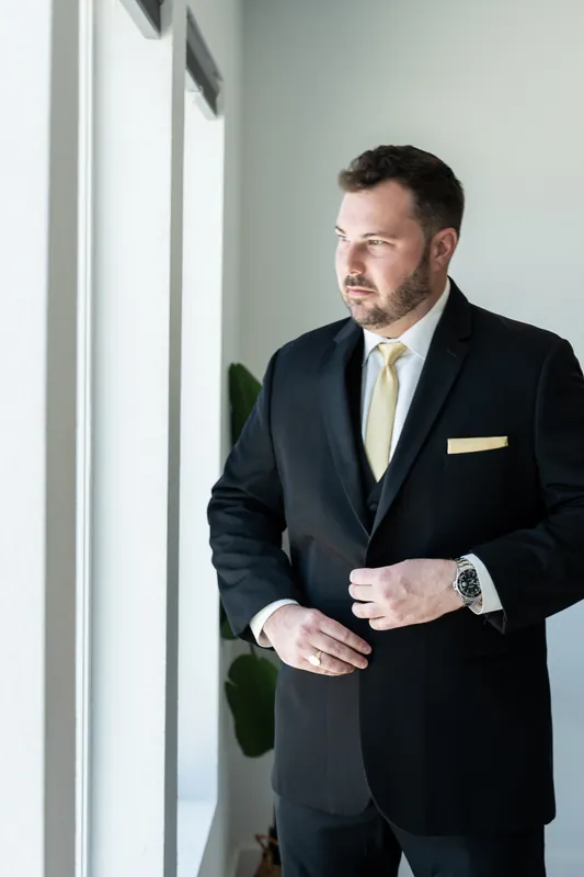 Stephen stands by a window in dark suit, gold tie, and pocket square, looking thoughtful.