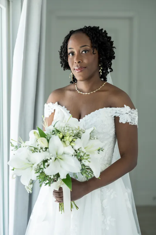 Malika in an off-shoulder white lace gown holds a white and green bouquet, smiling softly at the Airbnb in St. Augustine.