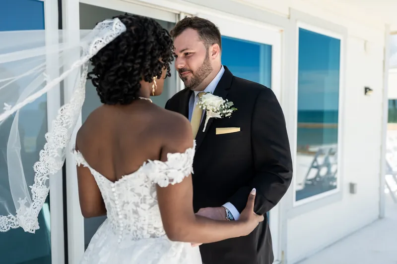 Stephen and Malika face each other indoors, exchanging words with ocean views visible through glass doors behind them.