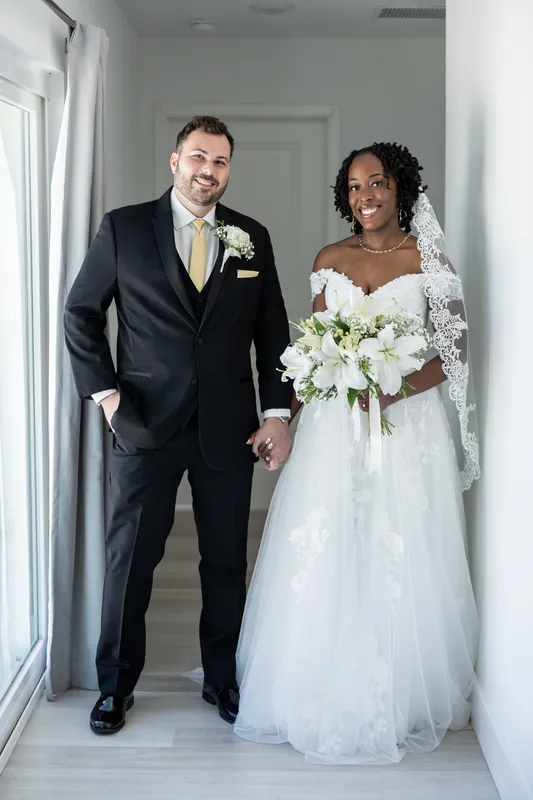 Stephen in a dark suit and Malika in her white gown walk down a bright hallway holding hands and bouquet.