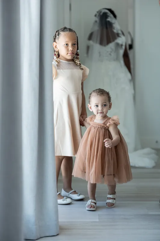Two children stand in a hallway—one in cream, one in dusty rose tulle—with a blurred figure behind them.