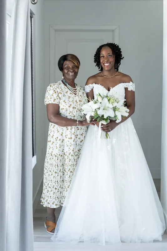 A woman in a cream patterned dress stands beside Malika, who holds her white bouquet, both framed in a doorway at the Airbnb.