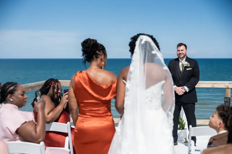 A person in an orange dress holds a camera while a couple exchanges vows on a seaside deck with guests watching.