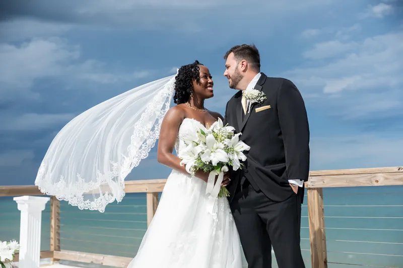 Stephen and Malika embrace on a beachfront deck, her veil billowing as they hold a white bouquet with the ocean behind them.