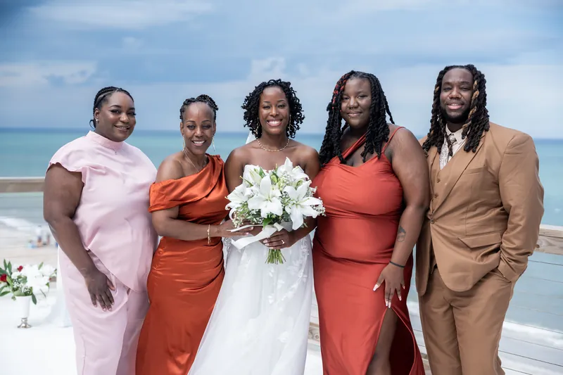 Five wedding guests in coral, pink, and tan stand together on a deck; the center figure holds a white bridal bouquet.
