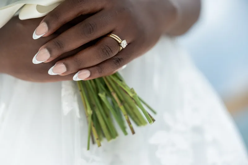 Close-up of two hands holding a green-stemmed white bridal bouquet, one hand wearing a wedding band.