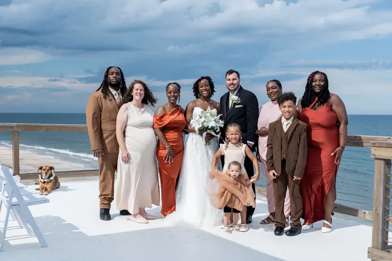 A wedding party of ten including a dog stands on a white deck overlooking the ocean in shades of coral, cream, and brown.