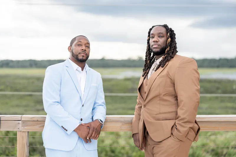 Two people stand on a wooden railing overlooking a wide river valley—one in a light blue suit, the other in a tan blazer.