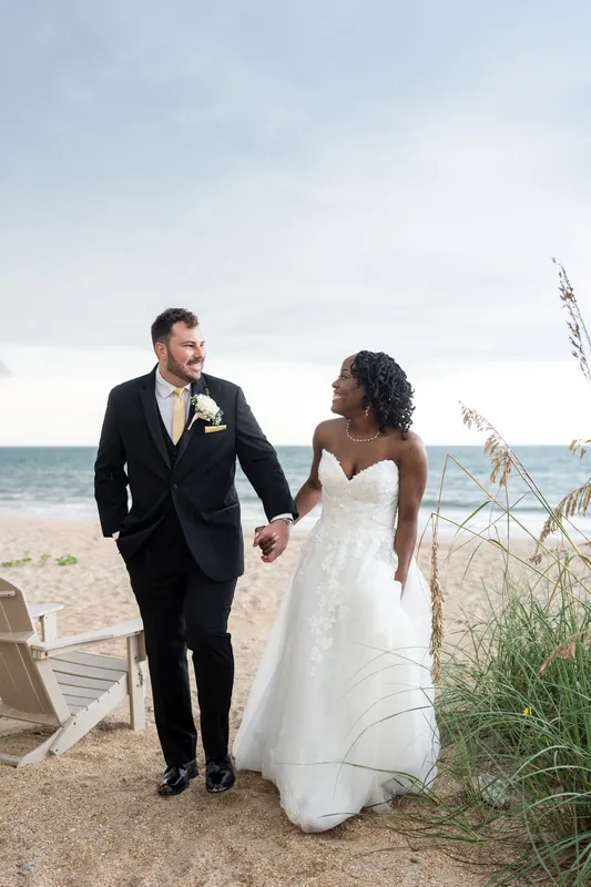 Stephen and Malika hold hands on a beach near dune grasses in their wedding attire, ocean behind them.