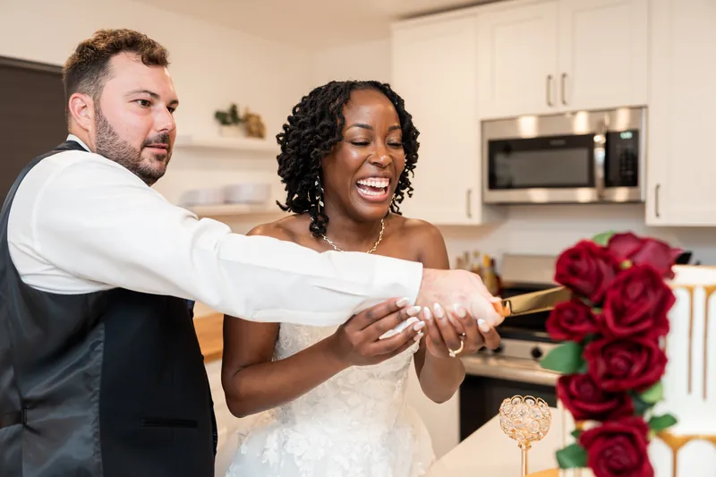 The couple laughs together while cutting their cake in a bright kitchen, deep red roses visible on the table.
