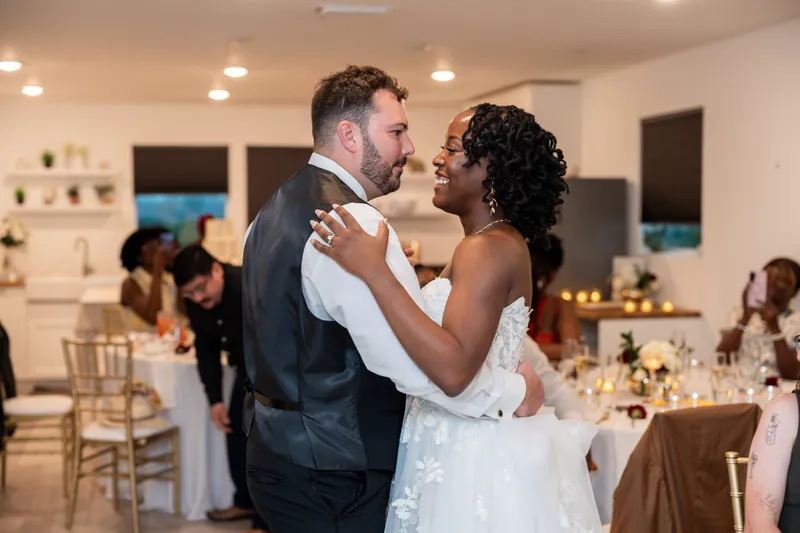 Stephen and Malika dance during their reception in a light-filled room, guests seated at tables in the background.