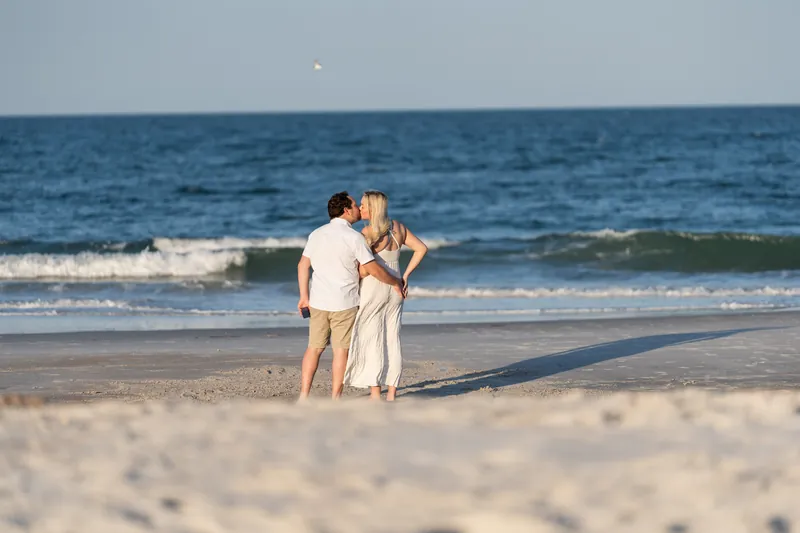 Steve kneels on the sand at Vilano Beach while Paige stands facing him in a cream linen dress, ocean and waves behind them.