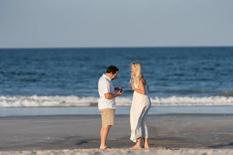 Steve holds an open ring box toward Paige, who looks down at it, both standing in shallow water at the beach's edge.