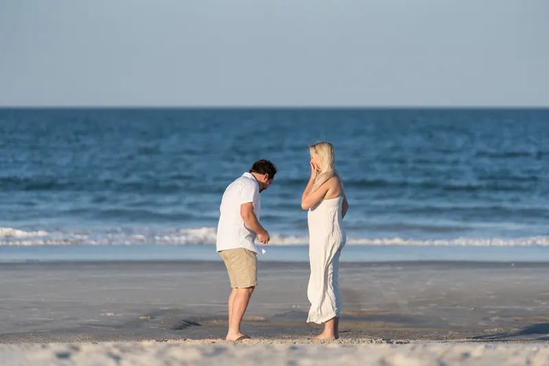 Steve presents the ring box to Paige as she reacts with her hand near her face, waves breaking behind them on the sand.