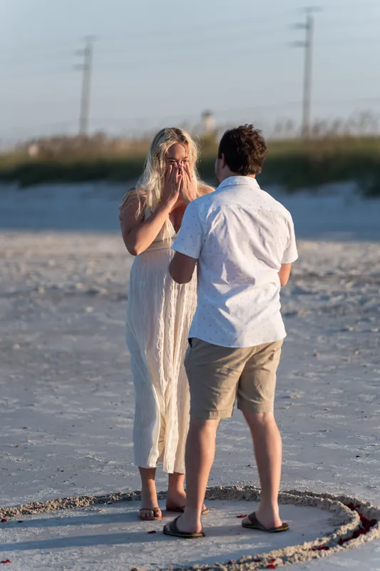 Paige touches her face in emotion while Steve stands beside her in the shallow water, a heart drawn in the sand around them.