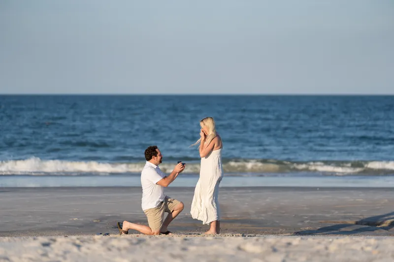 Steve kneels on one knee in the sand proposing to Paige, who stands in her cream dress with the ocean stretching behind them.