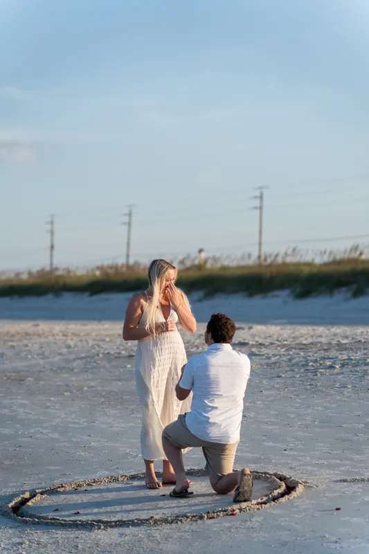 Steve kneels in shallow water as Paige stands before him in a cream dress, hands raised in surprise.