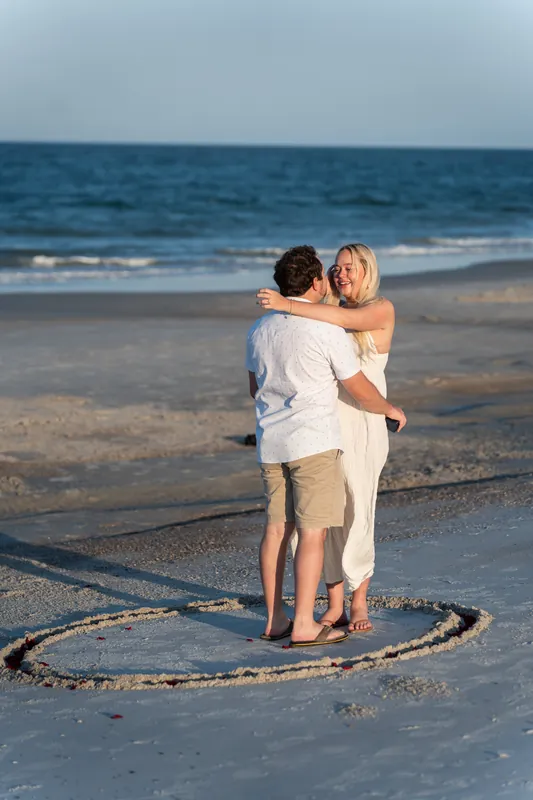 Steve embraces Paige from behind in the wet sand, a heart shape drawn around them on the beach.
